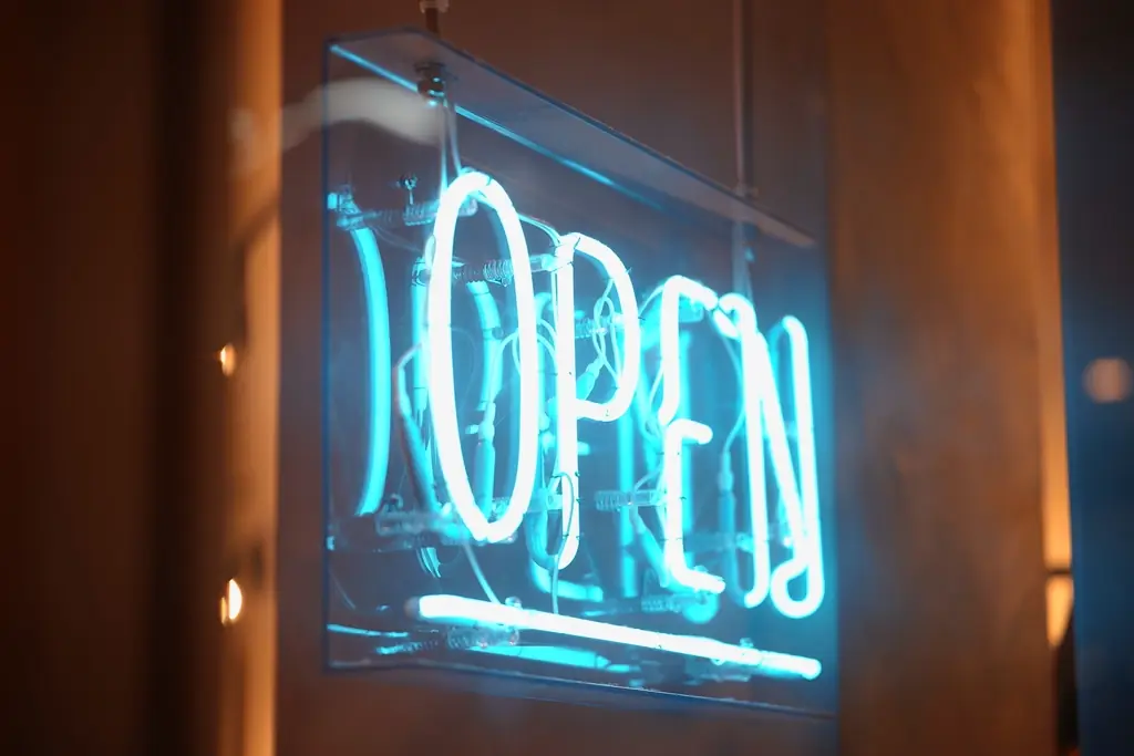 Letrero de neón cian con la palabra «OPEN» sobre un vidrio, con un fondo cálido desenfocado.