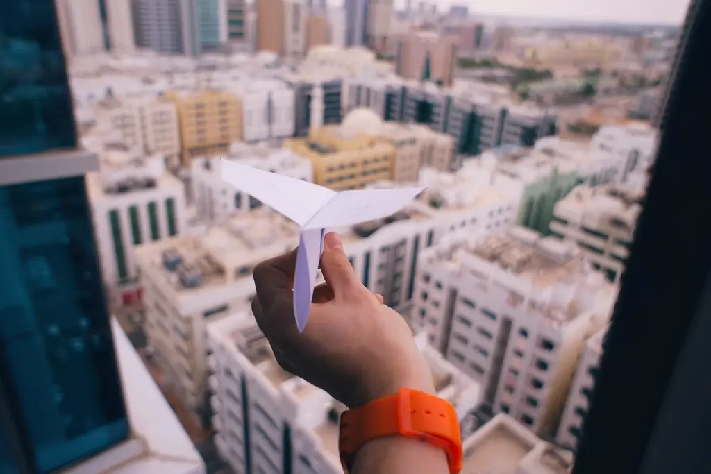 Una mano sostiene un avión de papel blanco sobre una ciudad vista desde lo alto.