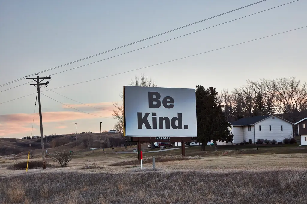 Un cartel publicitario blanco con el mensaje «Be Kind.» en un campo al atardecer, junto a postes de luz y una casa.