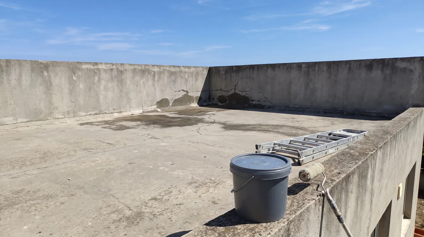 Azotea de concreto con muros altos, manchas oscuras de humedad en el piso y en la esquina del fondo, y grietas finas; en primer plano, sobre el remate del muro, un balde gris cerrado sin etiqueta, una escalera de aluminio plegada y un rodillo de pintura apoyado en el muro, bajo un cielo azul.