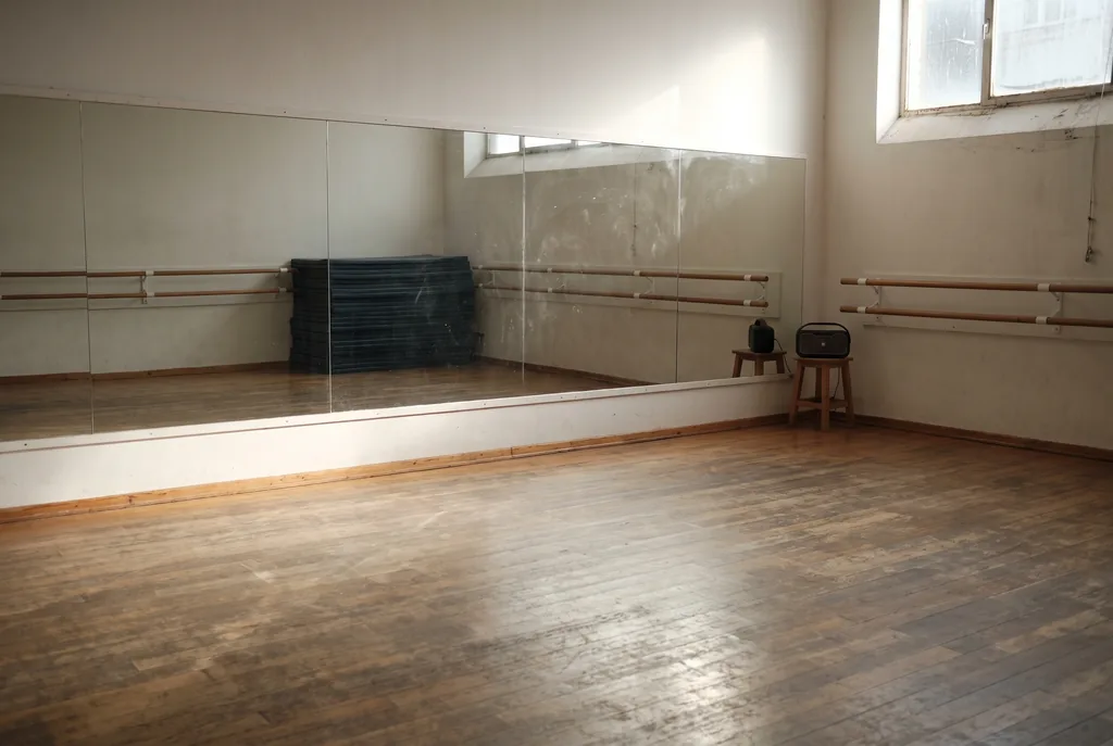 Academia de baile vacía por la mañana, con un espejo largo en la pared que refleja el salón, barras de ballet de madera a lo largo del espejo y de la pared lateral, un parlante portátil sobre un banco, una pila de colchonetas oscuras y el piso de madera gastado con luz de una ventana alta