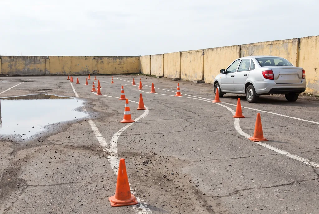 Patio de práctica de manejo con conos naranjas en zigzag sobre un piso de asfalto agrietado con líneas blancas gastadas, un auto plateado estacionado junto a un muro amarillento y un charco que refleja un cielo gris claro