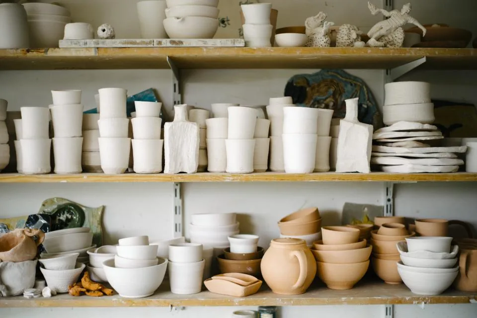 Repisas de un taller de cerámica con tazas, cuencos y piezas de arcilla blanca y de barro cocido.